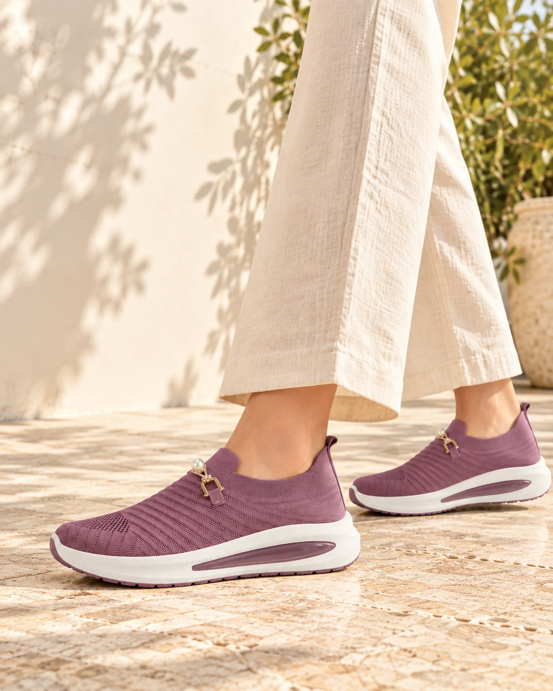 Mauve pearl-accent knit slip-ons worn with cream linen trousers on a sunlit terrace, photographed below the knees
